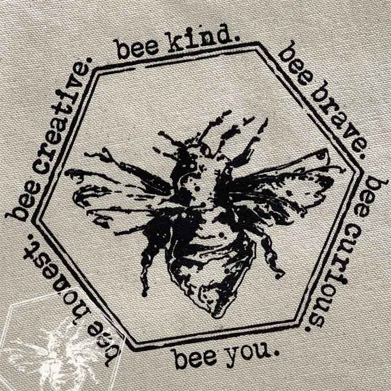 A close-up of a beige fabric surface printed with a black hexagonal bee illustration and decorative text around it, including phrases like 'bee kind.', 'bee brave.