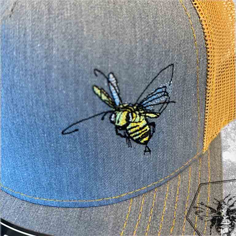 Close-up of a grey denim trucker hat with orange mesh back, featuring an embroidered bee design with detailed wings and yellow-and-black striped body on the front panel.