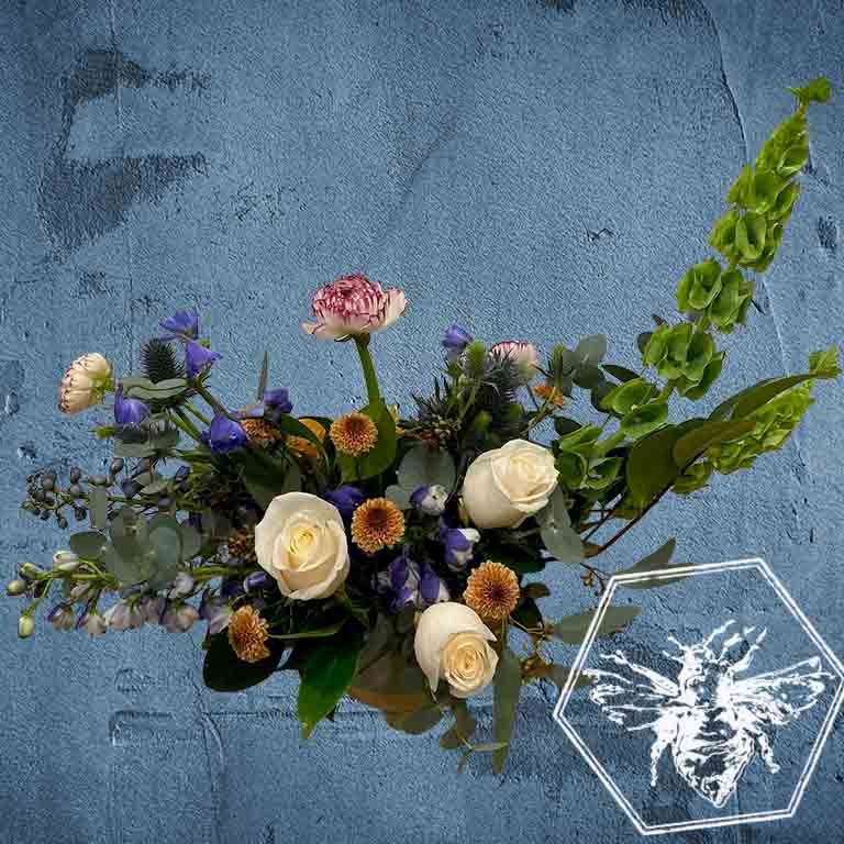 A modern, horizontally styled sympathy floral arrangement in a neutral ceramic pot, featuring a lush mix of premium white and soft pastel blossoms accented with green foliage and blue accents 