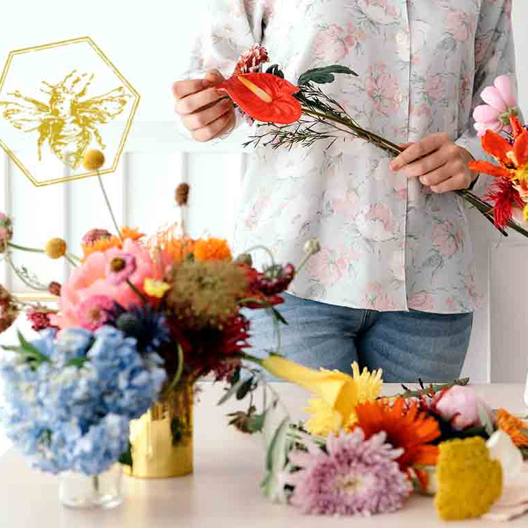 A person in a light floral print shirt and blue jeans stands behind a table filled with colorful flower arrangements in vases and loose stems, holding a red-orange flower. The scene is brightly lit with a clean white background.
