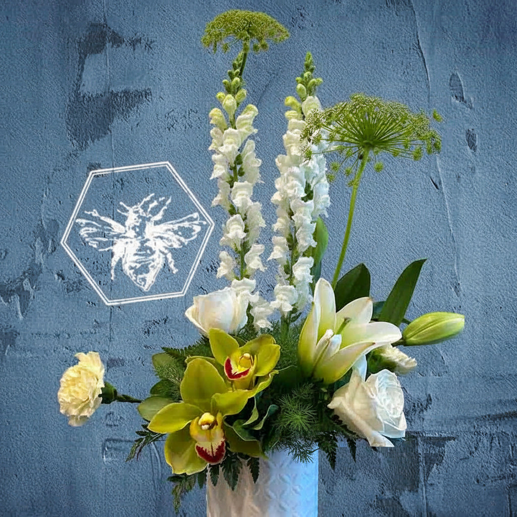 A modern, vertically styled sympathy floral arrangement in a neutral ceramic pot, featuring a lush mix of premium white and soft pastel blossoms accented with green foliage