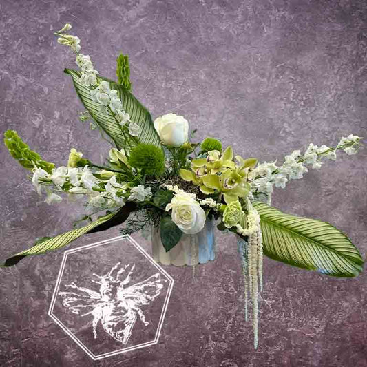 A modern, horizontally styled sympathy floral arrangement in a neutral ceramic pot, featuring a lush mix of premium white and soft pastel blossoms accented with cascading green foliage