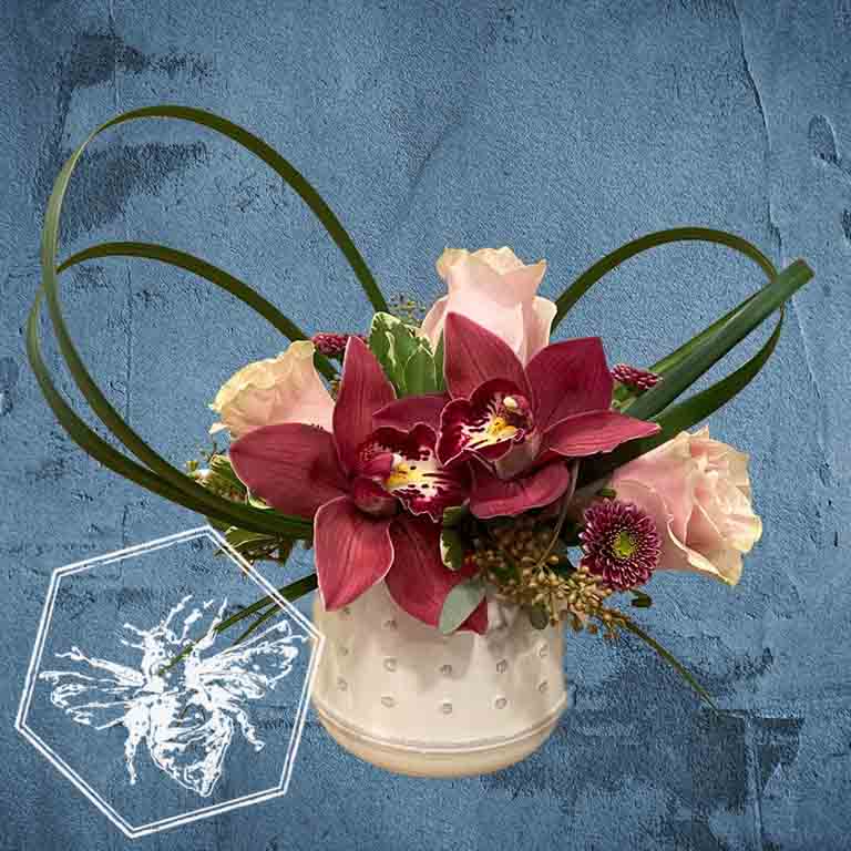 Romantic roses greenery, button mums and giant orchids nestled in lily grass accents in a white ceramic pot