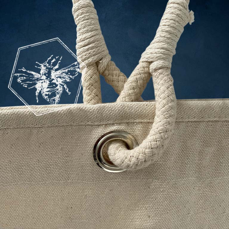 Close-up of a natural canvas tote bag with thick braided rope handles and a metal grommet, set against a dark blue background with a white bee logo.