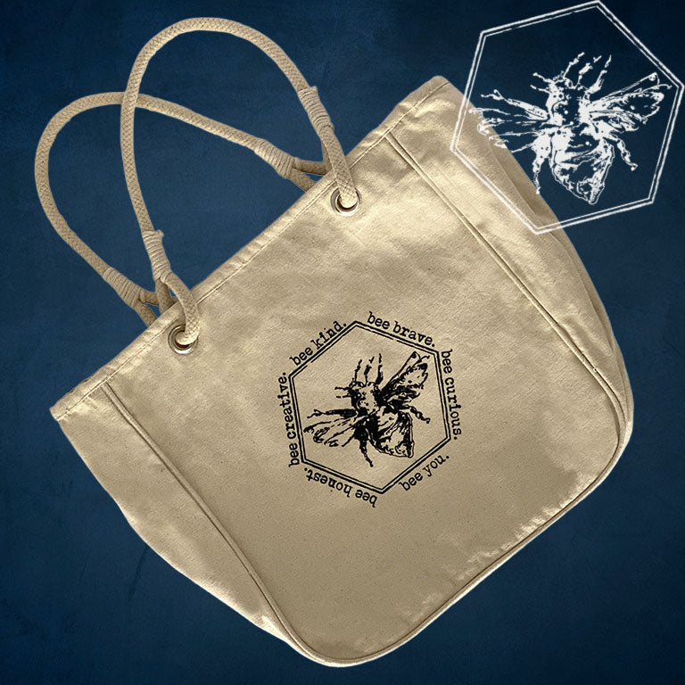 A beige canvas tote bag with rope handles, featuring a black printed bee graphic and the text 'bee brave' and 'bee creator' on the front, set against a dark blue textured background with a white hexagonal bee graphic overlay in the top right.
