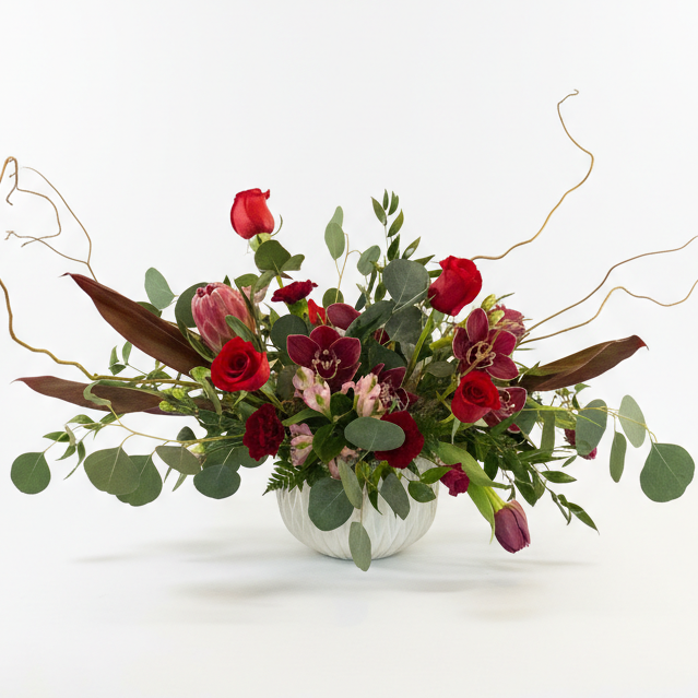All of the colors of love wrapped up in modern luxurious fresh floral creative brilliance featuring red roses, tropical protea, twig and silver dollar eucalyptus