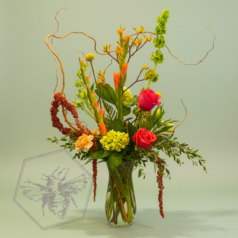 A bold, generous mix of vivid fresh flowers in a classic clear vase featuring white star, button mums, roses, alstroemeria, and greenery