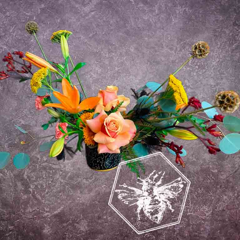 A modern, horizontally styled sympathy floral arrangement in a neutral ceramic pot, featuring a lush mix of premium colorful blossoms accented with green foliage