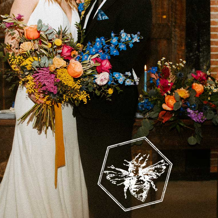 A bride in a white wedding dress and a groom in a black suit hold a large, colorful wildflower bouquet together in front of a brick wall. A matching floral arrangement sits on a table beside them, and a hexagonal bee-shaped decorative plaque is overlaid near the bottom center.